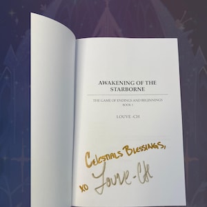 May include: An open book with the title "AWAKENING OF THE STARBORNE" and the author's name "LOUVE-CH" printed on the page. The book is signed with gold ink, reading "Celestial Blessings, xo Jouve-lfe."