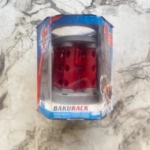 Peut inclure: Un ensemble de jouets Bakugan Bakurack dans son emballage d'origine. L'emballage est bleu et présente le logo Bakugan et des illustrations de personnages. Le Bakurack lui-même est rouge et gris, conçu pour contenir des figurines Bakugan. Le texte "BAKURACK" est visible.