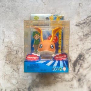 May include: A Patamon action figure in a clear plastic display case. The figure is orange and white with large ears and blue eyes. The packaging includes the word "PATAMON" and a cartoon illustration.