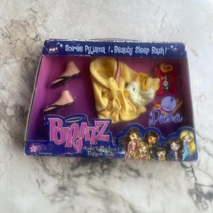 May include: A Bratz Mode Collection fashion pack with a yellow robe, slippers, and pajamas. The box reads "Soirée Pyjama ! Beauty Sleep Bash!" and features the Bratz logo and illustrations of the Bratz characters.