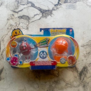 May include: A Pac-Man and the Ghostly Adventures toy set featuring two characters in a blister pack. One is red with a smiling face, and the other is orange. The packaging includes the text "Pac-Man" and "2 in 1".