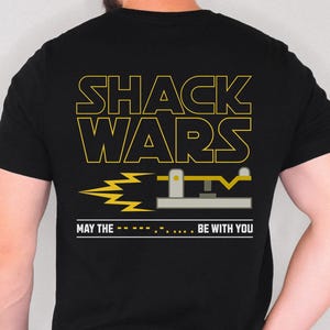 May include: Black t-shirt featuring the yellow text "SHACK WARS" and a graphic of a shack. The phrase "MAY THE... BE WITH YOU" is also printed on the shirt. The t-shirt is a crew neck and is being worn.