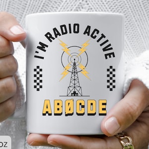 May include: White ceramic mug with the text "I'm Radio Active" in black, yellow, and white. The design includes a radio tower, lightning bolts, and the letters "ABØCDE". The mug is 11oz.