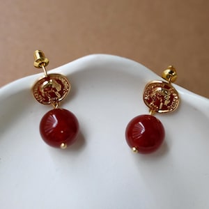 Pode incluir: Um par de brincos de tarracha dourados com um pingente circular estilo moeda com um design de relevo de perfil, suspenso sobre uma conta redonda e lisa de cor vermelho profundo. Incluem fechos de pressão dourados para o lóbulo.