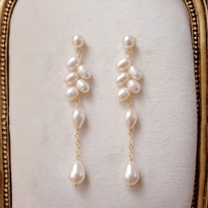 Può includere: Eleganti orecchini pendenti con grappoli di perle bianche sospese da delicate catene d'oro. Gli orecchini hanno una perla a forma di goccia nella parte inferiore, aggiungendo un tocco di raffinatezza. Gli orecchini sono su uno sfondo bianco sporco.