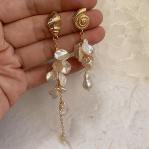Può includere: Orecchini a conchiglia dorati con pendenti iridescenti a forma di petalo e una singola perla. Gli orecchini hanno un design unico e asimmetrico, con uno caratterizzato da una catena di perle più lunga. Eleganti orecchini.