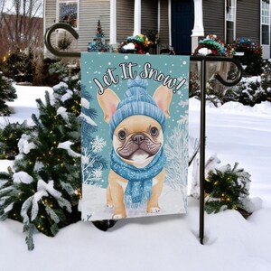 May include: A garden flag featuring a tan French Bulldog wearing a blue knit hat and scarf, with the text "Let It Snow!" against a light blue background with snowflakes. The flag is displayed outdoors in a snowy environment.