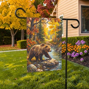 May include: A rectangular garden flag hanging on a black metal stand, featuring a colorful illustration of a brown bear standing on a rock in a stream. The scene depicts a forest with autumn foliage in shades of yellow, orange, and red.