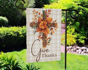 May include: A garden flag with a cross design, adorned with autumn leaves in shades of orange and brown. The flag features the words "Give Thanks" in a script font, set against a beige plaid background. The flag is displayed on a black metal stand.