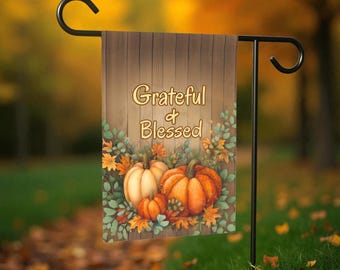 May include: A garden flag with the words "Grateful & Blessed" in gold lettering. The flag features a wooden background with pumpkins, leaves, and foliage in shades of orange, green, and brown. The flag is hanging on a black metal stand.