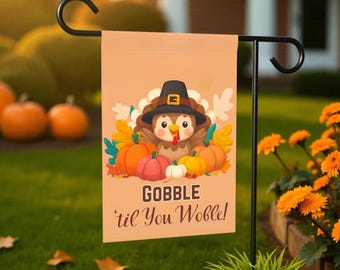 May include: A tan garden flag featuring a cartoon turkey wearing a pilgrim hat, surrounded by pumpkins and fall foliage. The flag reads "GOBBLE 'til You Wobble!" in a playful font, perfect for Thanksgiving.