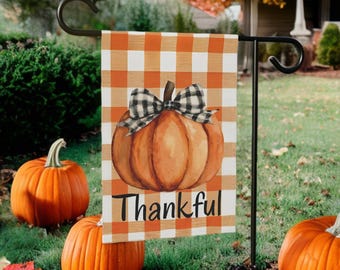 Può includere: Una bandiera da giardino a tema autunnale con un design di zucca e la scritta "Thankful" in nero. La bandiera ha uno sfondo a quadretti arancione e bianco. Tre zucche arancioni sono in primo piano.