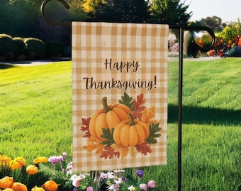 Könnte beinhalten: Eine Gartenflagge mit einem karierten Muster in Beige und Weiß. Die Flagge zeigt den Text "Happy Thanksgiving!" über einer Illustration von zwei orangefarbenen Kürbissen und Herbstblättern. Die Flagge befindet sich auf einem schwarzen Metallständer in einem Garten.