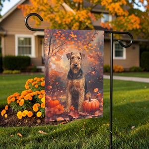 May include: A rectangular garden flag featuring an Airedale Terrier sitting amidst fallen autumn leaves and orange pumpkins. The scene is set against a backdrop of trees with golden foliage, displayed on a black metal stand in a grassy yard.