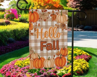 May include: A garden flag with a fall theme. The flag features a tan and white checkered background with orange pumpkins and fall leaves. The words "Hello Fall" are printed in orange and brown. The flag is displayed on a black metal stand in a garden setting.