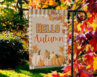May include: A garden flag with the words "HELLO Autumn" in orange and brown. The flag features a plaid background with pumpkins in a basket. The flag is displayed on a black metal stand, surrounded by fall foliage.