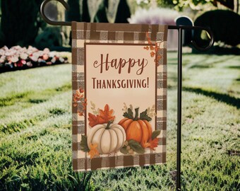 Könnte beinhalten: Eine Thanksgiving-Gartenflagge mit einem braunen Karomuster-Rand und dem Schriftzug "Happy Thanksgiving!". Die Flagge zeigt einen weißen und orangefarbenen Kürbis mit Herbstblättern. Sie ist auf einem schwarzen Metallständer in einem grasbewachsenen Garten befestigt.