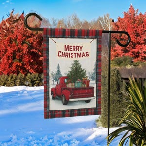 May include: A festive garden flag with a red vintage truck laden with a Christmas tree and presents. The flag has a red and black plaid border and the words "MERRY CHRISTMAS". The flag is displayed on a black metal stand in a snowy garden setting.
