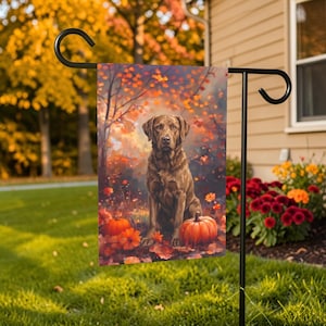 May include: A rectangular garden flag featuring a brown Labrador retriever sitting in a forest setting with orange autumn leaves, scattered foliage, and two orange pumpkins on the ground, suspended from a black metal garden flag stand.