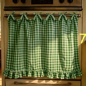 Op de afbeelding: Een groen en wit geblokt keukengordijn met een gefranjerde onderrand en strikken aan de bovenkant. Het gordijn hangt voor een vintage oven. Het algemene thema is boerderijkeukeninrichting.