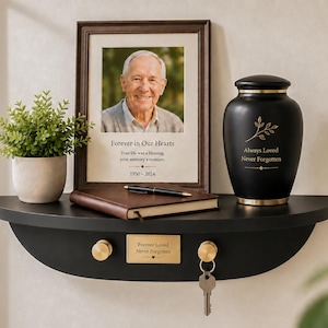 Könnte beinhalten: Ein schwarzes, halbkreisförmiges Wandregal mit zwei goldenen Haken, einer goldenen Plakette mit der Aufschrift "Forever Loved Never Forgotten", einem gerahmten Foto eines älteren Mannes, einer schwarzen Urne mit goldenem Text, einem braunen Notizbuch, einem Stift und einer kleinen grünen Pflanze.