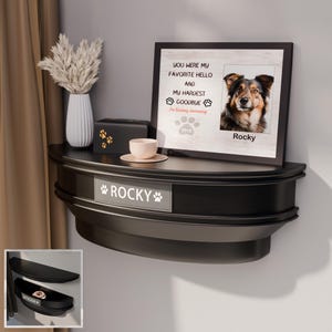 May include: A black pet memorial shelf with the name "ROCKY" and paw prints. The shelf holds a framed photo of a dog, a small box, a cup and saucer, and a vase with dried flowers. The photo reads "YOU WERE MY FAVORITE HELLO AND MY HARDEST GOODBYE".
