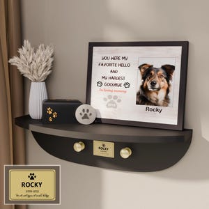 May include: A memorial shelf with a framed photo of a dog named Rocky, a black urn with paw prints, and a decorative paw print stone. The photo frame reads "You were my favorite hello and my hardest goodbye."