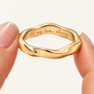 Peut inclure: Une bague dorée avec une surface de bande irrégulière et ondulée. L'intérieur de la bague présente le texte gravé en cursive "All my love, always". Le métal a une finition polie et réfléchissante qui capte la lumière sur ses bords incurvés.