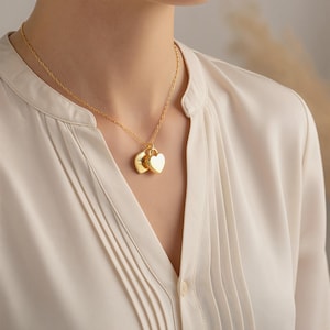 Pu&ograve; includere: Una collana color oro con una catenina sottile e due piccoli ciondoli a forma di cuore, lisci e lucenti. I ciondoli presentano una superficie levigata e riflettente, sospesi insieme da un semplice anello circolare sulla catena.