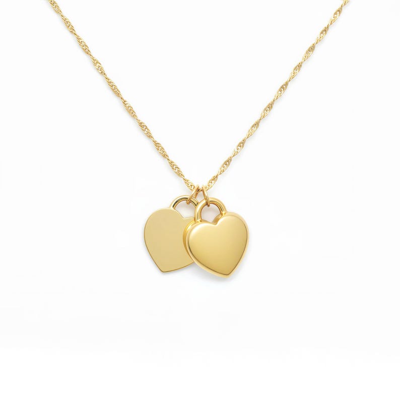 Pu&ograve; includere: Una collana color oro caratterizzata da una sottile catena ritorta e due ciondoli lisci a forma di cuore che pendono affiancati. I ciondoli hanno una superficie lucida e riflettente e sono fissati alla catena tramite piccoli anellini.
