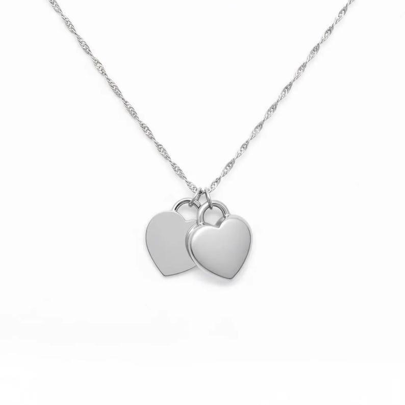 Pu&ograve; includere: Una collana color argento con una sottile catenina intrecciata e due ciondoli a forma di cuore, lisci e lucidi, che pendono affiancati. La superficie metallica &egrave; riflettente e semplice, caratterizzata da un design essenziale senza incisioni.