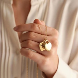 Pu&ograve; includere: Una collana color oro con due ciondoli a forma di cuore sospesi a una catenina sottile. Un ciondolo a forma di cuore &egrave; liscio e lucido, mentre l'altro ciondolo a forma di cuore presenta un testo inciso visibile sulla superficie metallica.