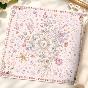 May include: A square light pink game mat featuring a central compass rose surrounded by intricate illustrations of seashells, starfish, coral, and swirling ocean motifs, accompanied by a matching beige fabric drawstring storage bag.