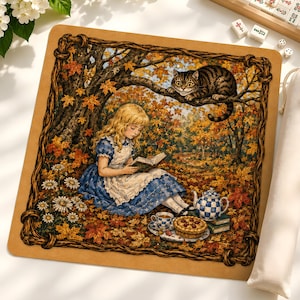 May include: A square mat featuring a detailed illustration of a blonde person in a blue dress reading a book under a tree with autumn leaves. A striped cat sits on a branch above, while a tea set and a pie are placed on the ground nearby.