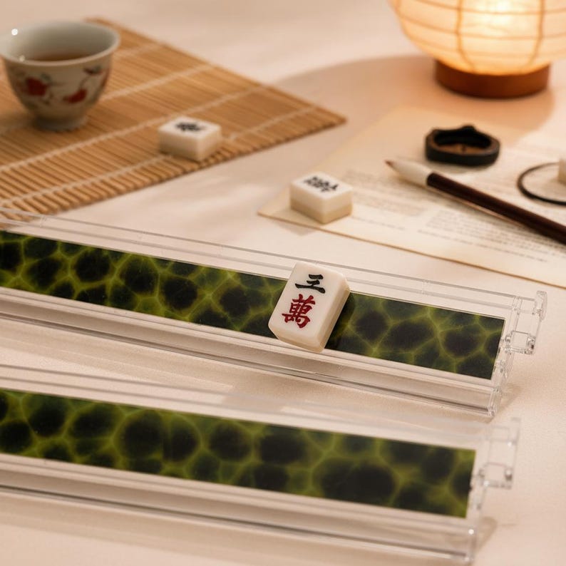 May include: Two clear acrylic mahjong tile racks featuring a green and black mottled pattern. One rack holds a single white mahjong tile with the black and red Chinese character for three of wan. The racks are rectangular with raised edges.