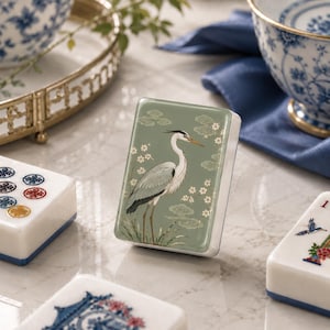 Può includere: Una tessera rettangolare da mahjong con una superficie verde salvia e un'illustrazione dettagliata di un airone grigio in piedi tra fiori di ciliegio bianchi stilizzati e nuvole. La tessera ha una finitura liscia e lucida e base bianca.
