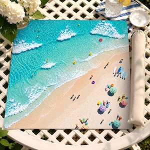 May include: A square beach mat featuring an aerial view of a turquoise ocean with white waves, a sandy shore, and colorful beach umbrellas, surfboards, and swimmers. The mat is displayed alongside a matching beige fabric carrying case.