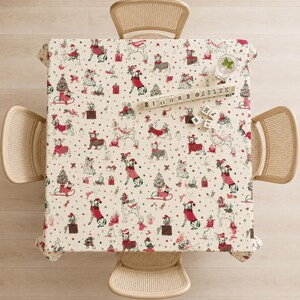 Könnte beinhalten: Eine quadratische Tischdecke mit cremefarbenem Hintergrund und einem wiederkehrenden Muster aus Hunden in roten und grünen Weihnachtspullovern und Mützen, durchsetzt mit kleinen Tannenbäumen, Geschenken und Sternen, mit einem Spielsteinhalter aus Holz.
