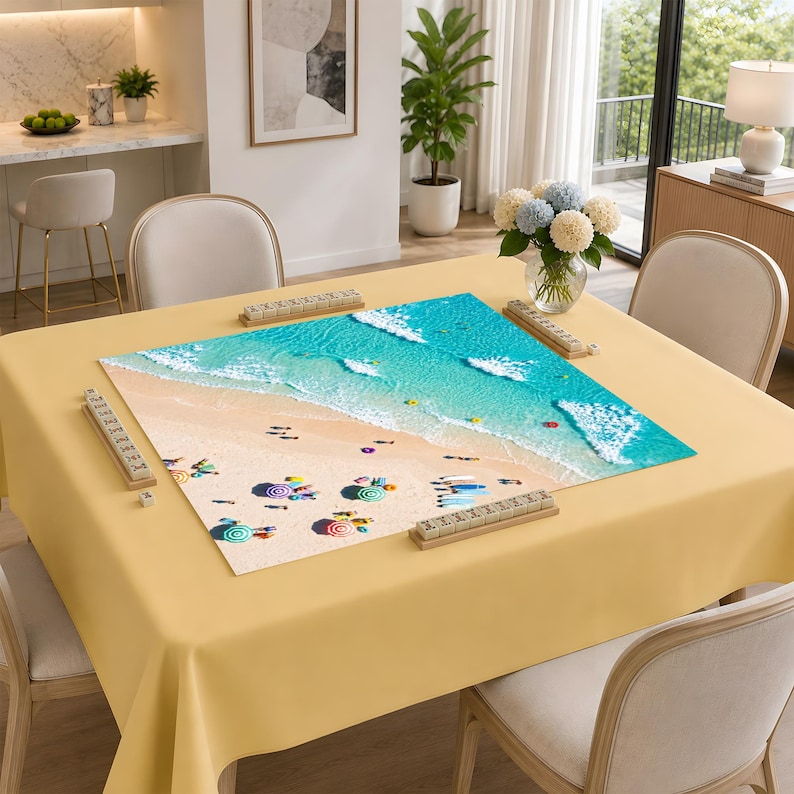 May include: A square game board featuring a high-angle view of a sandy beach with turquoise ocean waves, colorful beach umbrellas, and small figures. The board is placed on a solid light yellow tablecloth, surrounded by four wooden tile racks.