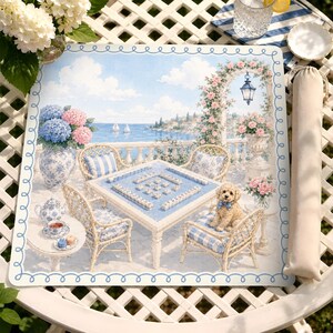 May include: A square mahjong mat featuring a coastal terrace scene with a table set for play, wicker chairs, a small light-colored dog, blue and white floral accents, and a blue scalloped border, accompanied by a matching beige fabric storage bag.