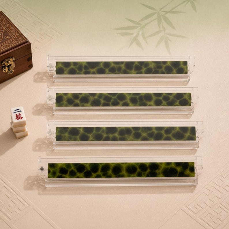 May include: Four rectangular mahjong tile racks made of clear acrylic, each featuring a dark green and black mottled pattern along the front face. The racks are arranged horizontally in a stack, accompanied by a small wooden box and three mahjong tiles.