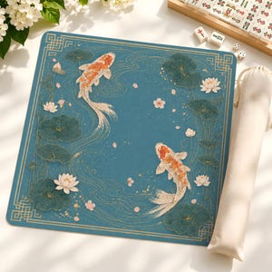 May include: A square blue mat featuring two orange and white koi fish swimming among dark green lily pads and white lotus flowers. The design includes gold line art of water ripples and a geometric border, accompanied by a beige fabric storage bag.