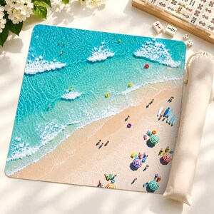 May include: A square mouse pad featuring an aerial view of a sandy beach with turquoise ocean water, white crashing waves, and colorful beach umbrellas. Several small figures are scattered across the sand and in the water, accompanied by a beige drawstring bag.