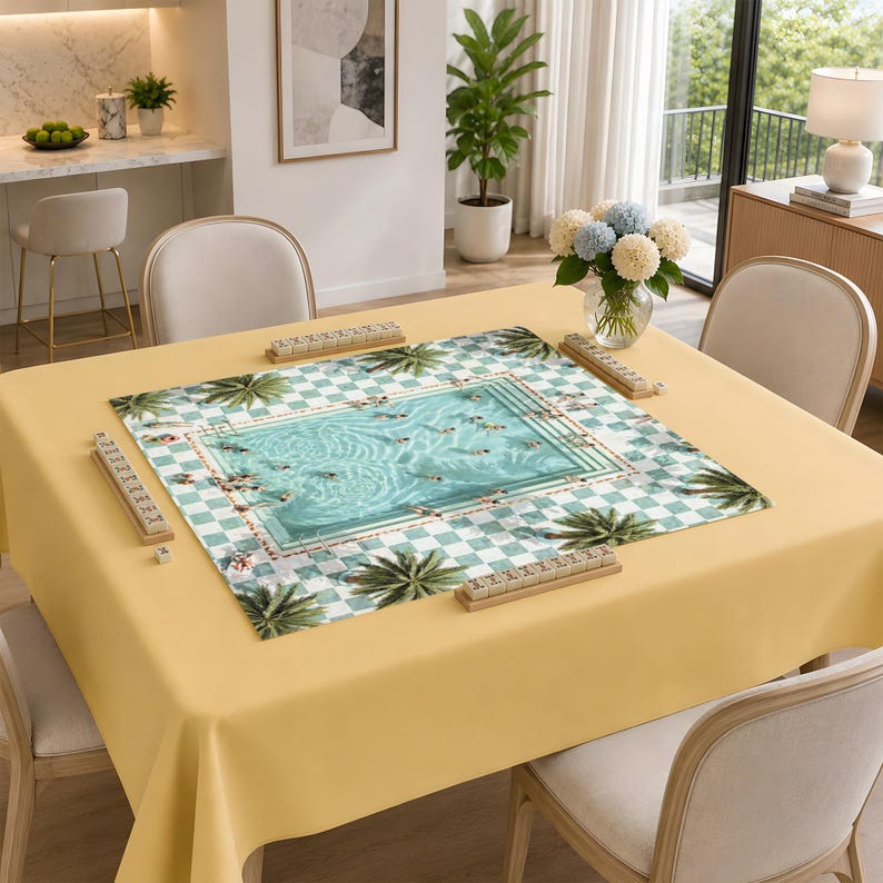 May include: A square mahjong mat featuring a light blue swimming pool graphic surrounded by a white and green checkered border with palm tree illustrations. Four wooden tile racks are positioned on the edges of the mat on a yellow tablecloth.