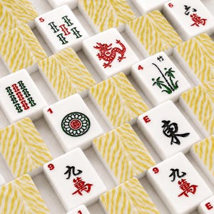 May include: A collection of Mahjong tiles featuring white faces with various red, green, and black symbols, including a red dragon, bamboo, and numerical characters. The backs of the tiles display a textured yellow and white tiger stripe pattern.