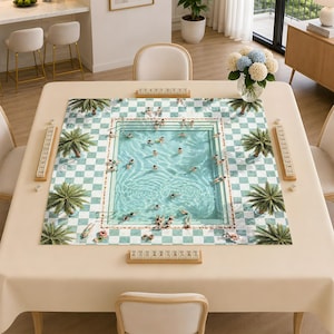 May include: A square mahjong mat featuring a printed design of a swimming pool surrounded by a green and white checkered patio with palm trees. Four wooden tile racks are positioned along the edges of the mat on a beige table surface.