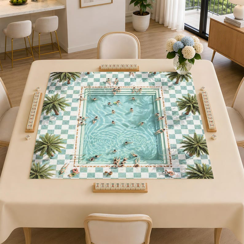 May include: A square mahjong mat featuring a printed design of a swimming pool surrounded by a green and white checkered patio with palm trees. Four wooden tile racks are positioned along the edges of the mat on a beige table surface.