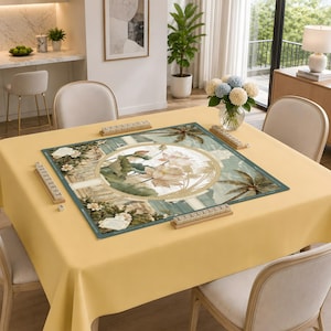 Puede incluir: Una alfombrilla de mahjong cuadrada sobre un mantel amarillo claro. La alfombrilla presenta una ilustraci&oacute;n circular central de flores de loto rodeadas de palmeras y columnas arquitect&oacute;nicas. Cuatro soportes de madera est&aacute;n en los bordes.