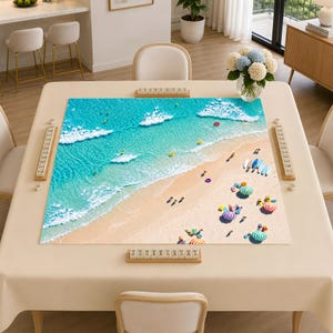 May include: A square mahjong table covered with a light beige cloth, featuring a large, high-angle aerial print of a turquoise ocean beach scene. The table includes four wooden tile racks placed along the edges, each holding mahjong tiles.