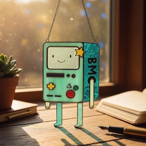 Puede incluir: Un personaje BMO de Adventure Time en vidriera, en tonos verdes, azules y amarillos. El personaje tiene una cara sonriente, una estrella y las letras "BMC" en un lado. Cuelga de una cadena.